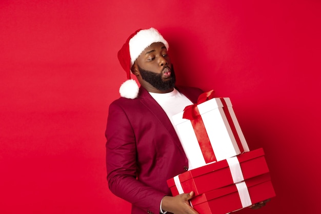 Christmas, New Year and shopping concept. Funny african american man in santa hat carry heavy xmas presents, holding gifts, standing over red background