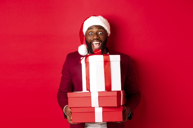 Christmas, New Year and shopping concept. Happy Black man in santa hat and blazer holding xmas presents, bring gifts and smiling, standing against red background.