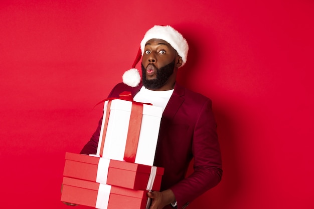 Christmas, New Year and shopping concept. Happy Black man in santa hat and blazer holding xmas presents, bring gifts and smiling, standing against red background