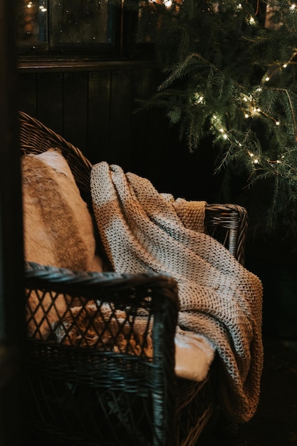 Christmas and new year time cozy armchair or wicker easy chair with a comfy knitted plaid and a fur pad pillow near a decorated christmas tree with glowing light winter holiday season at home