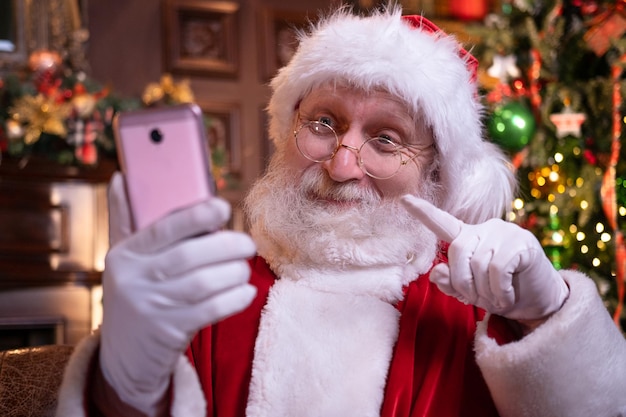 Christmas online congratulations from Santa Santa Claus using mobile phone for distance Christmas call people and kids Sitting near Christmas tree at home Happy New Year Covid 2021