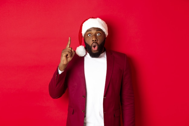 Christmas, party and holidays concept. Excited Black man in santa hat having an idea, raising finger up, suggesting plan, standing against red background