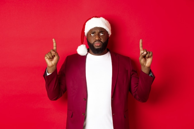 Christmas, party and holidays concept. Upset african-american guy in santa hat complaining, pointing fingers up and frowning disappointed, standing against red background