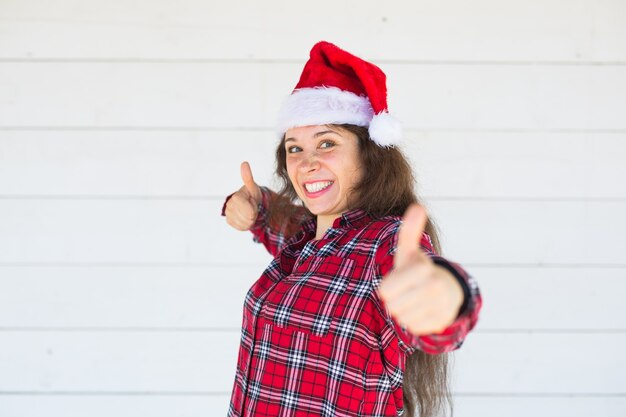 Christmas and people concept - joyful young woman in christmas hat showing thumbs up on white surface