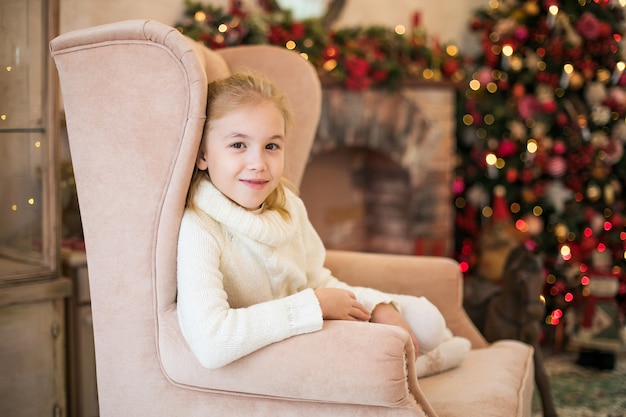 Christmas portrait of happy blonde child girl is sitting at home, decorated tree, presents and lights