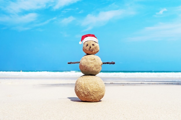 Christmas positive Sandy Snowman in red Santa Claus hat at ocean sunset beach. New Year vacation discounts in hot countries destinations concept