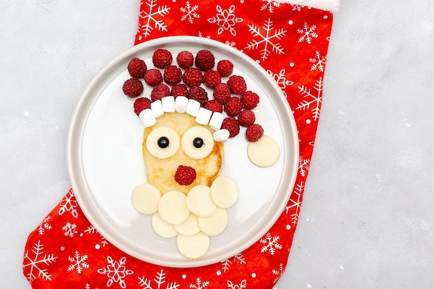 Christmas Santa Claus face shaped pancake with sweet fresh raspberry berry and cheese on plate on gray background for kids baby children breakfast dinner xmas food with new year decorations close up