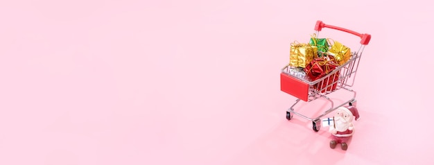 Christmas shopping concept mini red shop cart trolley with Santa Claus toy and gift box isolated on pale pink background blank copy space close up