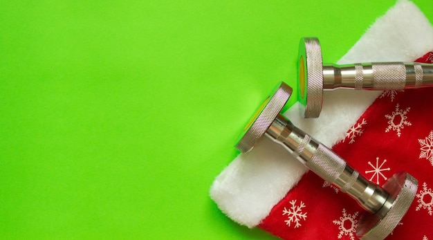 Christmas sport flatlay composition with dumbbells and christmas hat on green background.