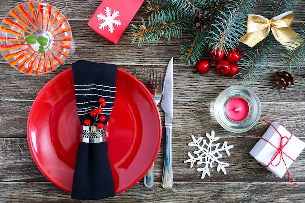 Christmas table setting. Red plate, fork, knife, martini glass, candle, napkin, gifts branch of a Christmas tree on a dark wooden background. Copy space. Christmas Xmas New Year holiday background.