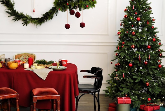 Christmas table setting On the table covered with a red tablecloth are plates cups and festive decor Christmas tree stands next to the table