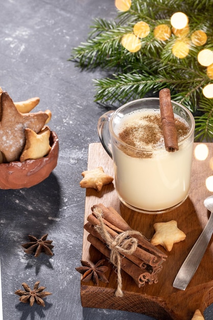 Christmas traditional drink eggnog in glass cup, heart shape cookies on wooden kitchen board and fir branches with bokeh lights on grey background. Cozy home Christmas. Vertical orientation.