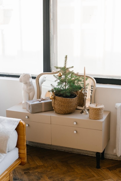 A Christmas tree and a gift box in a wicker basket stands on a light chest of drawers in the living room or bedroom in the interior of the house