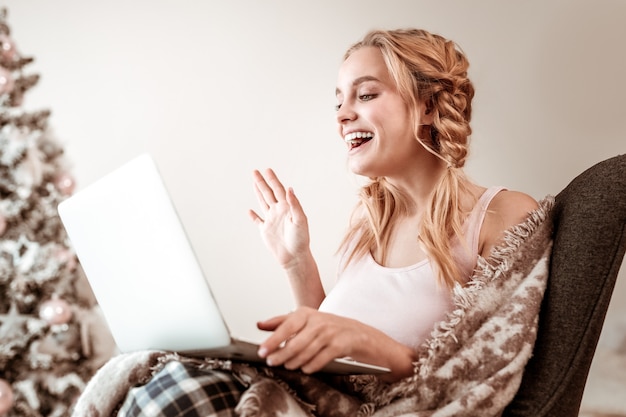 Christmas tree behind. Laughing blonde woman with two braids waving her hand while having online conference