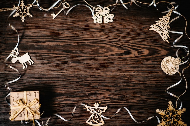 Christmas tree ornaments, gift, box with ribbons, snowflakes, bells, deer, angel on a brown, structural wood background. flat lay. top view, frame with space for text. happy holidays.