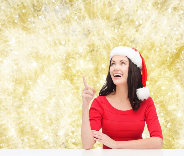 christmas, winter, holidays, happiness and people concept - smiling woman in santa helper hat pointing finger over yellow lights background