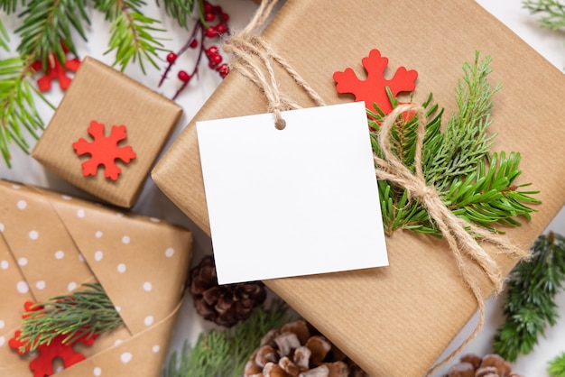 Christmas wrapped present with square paper gift tag on a white table with fir tree branches and decorations top view. Rustic winter composition with blank Gift tag Mockup, copy space