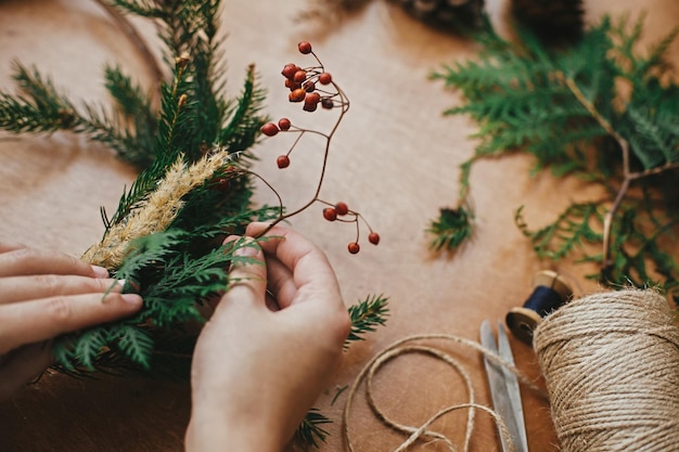 Photo christmas wreath workshop hands holding berries fir branches pine cones thread scissors on wooden table making rustic christmas wreath authentic rural wreath