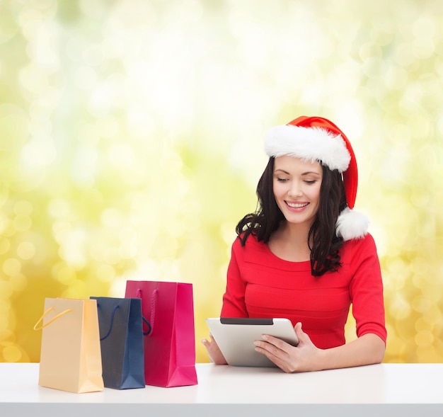 christmas, x-mas, online shopping and electronics concept - smiling woman in santa helper hat with shopping bags and tablet pc computer