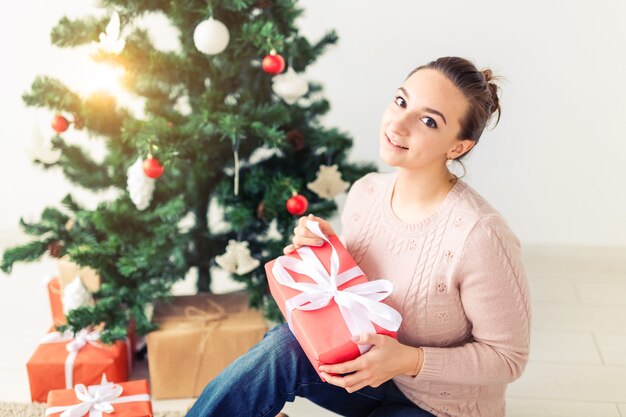 Christmas, x-mas, winter, happiness concept - girl opens a gift against the background of the Christmas tree. Happy young woman celebrating Christmas