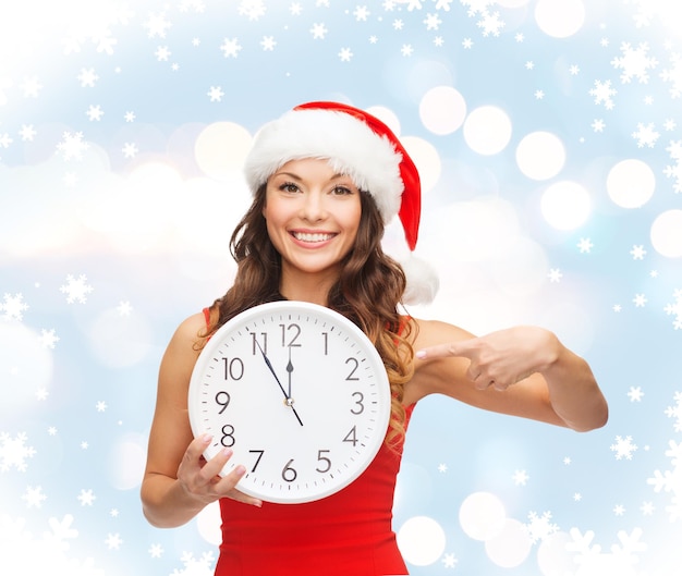 christmas, x-mas, winter, happiness concept - smiling woman in santa helper hat with clock showing 12