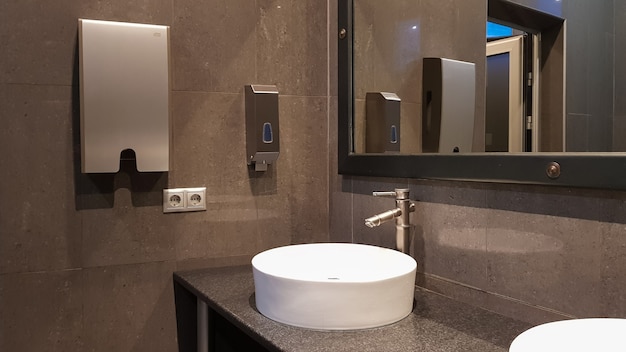 Chrome faucets with white round washbasins in a public toilet with a large mirror and gray walls, modern interior of a public toilet.