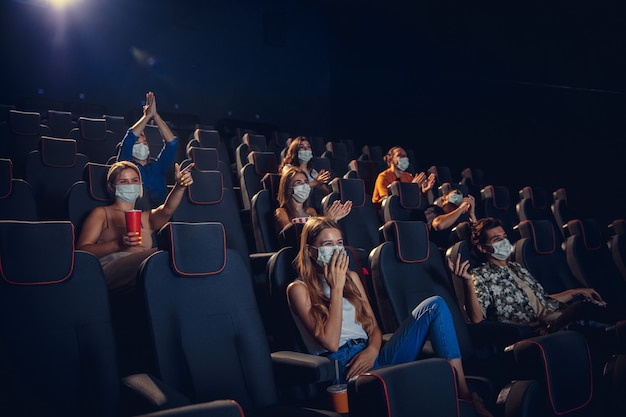 Cinema in quarantine. Coronavirus pandemic safety rules, social distance during movie watching. Men, women in protective face mask sitting in a rows of auditorium. Leisure time, youth culture concept.