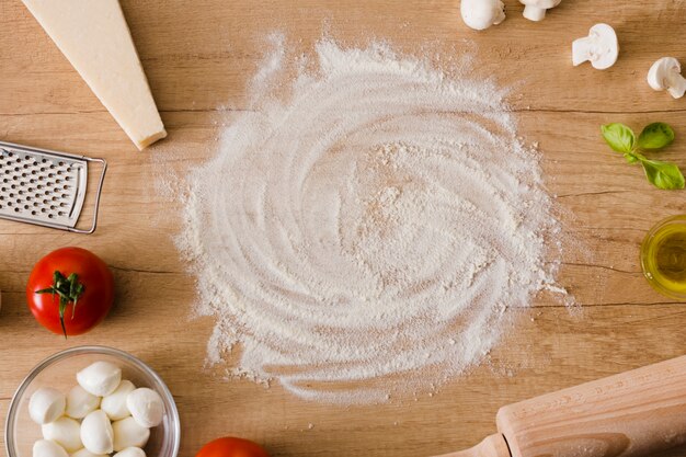 Circular flour surrounded with cheese block; mozzarella cheese; mushroom; basil and tomatoes on wooden table