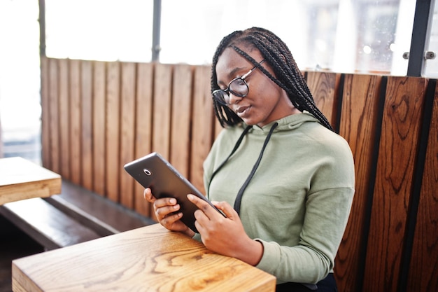 City portrait of positive young dark skinned female wearing green hoody and eyeglasses sitting with tablet