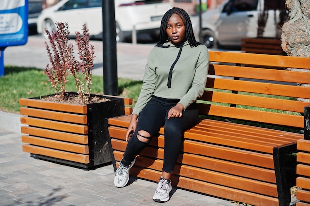 City portrait of positive young dark skinned female wearing green hoody sitting on bench.