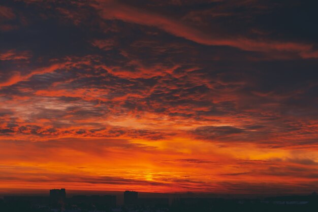 Cityscape with fiery blood vampire dawn. Amazing warm dramatic cloudy sky above dark silhouettes of city buildings. Orange sunlight. Atmospheric background of sunrise in overcast weather. Copy space.