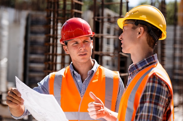 Civil engineer and construction manager in orange work vests and hard helmets explore construction documentation on the building site near the steel frames . .