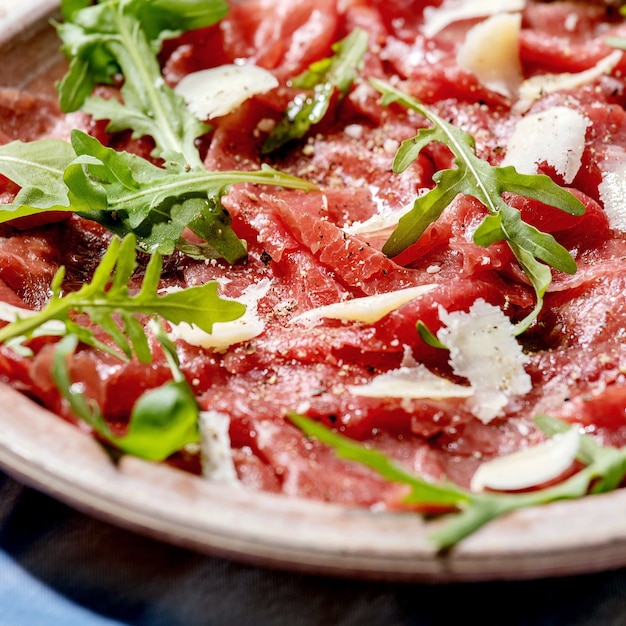 Classic beef carpaccio with cheese and arugula on ceramic plate over white and blue tablecloth. Square image