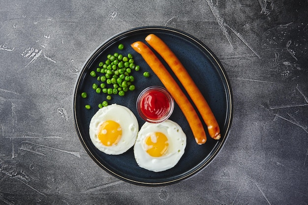 Classic breakfast of fried eggs sausages and green peas on dish on gray background