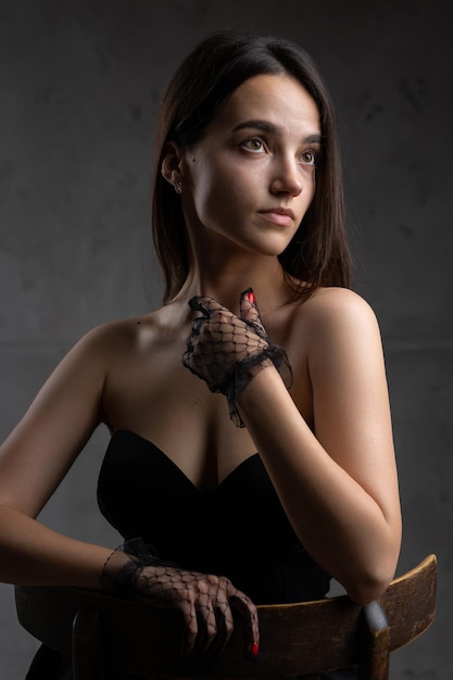 Classic dark studio portrait of a young brunette woman in black clothes who is sitting on a chair