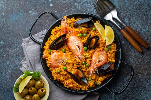 Classic dish of Spain, seafood paella in traditional pan on rustic blue concrete background top view. Spanish paella with shrimps, clamps, mussels, green peas and fresh lemon wedges from above