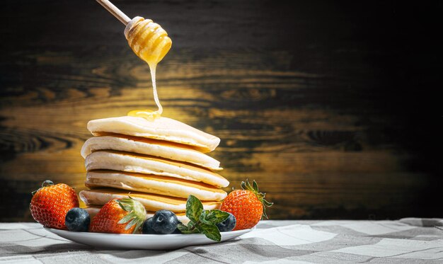 On classic homemade pancakes with berries on a white saucer on a dark wooden background in a rustic style pours delicious honey.