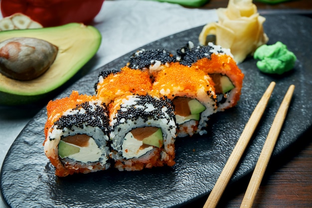 Classic japanese sushi rolls with salmon, avocado, cream cheese and caviar. California roll on a black plate with chopsticks. Japanese cuisine. Healthy seafood