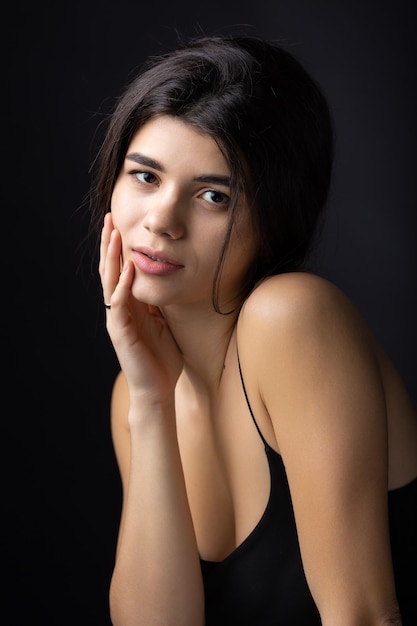 Classic studio portrait of a young brunette dressed in a black top who is sitting on a chair against a black background