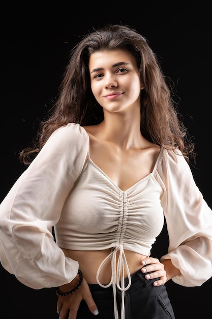 Classic studio portrait of a young brunette in a white loose top who is sitting on a chair against a black background