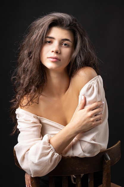 Classic studio portrait of a young brunette in a white loose top who is sitting on a chair against a black background