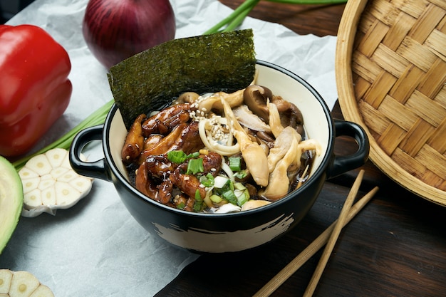 Classical Asian soup with udon noodles, nori, shiitake mushrooms, and wok-fried chicken in black bowl on wooden table with copy space. Top view, flat lay food