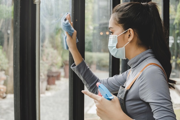 Clean asian waitress staff wearing protection face mask in apron cleaning window with disinfectant spray protect infection coronavirus covid19 in cafe coffee shop restaurant hotel service concept