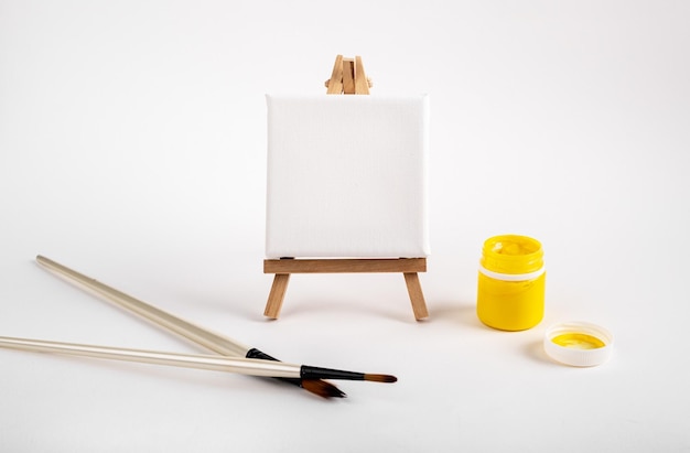 Photo clean canvas blank mockup for painting drawing on wood tripod easel paint brush and yellow gouache in open jar