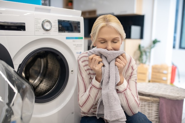 Clean clothes. Housewife in striped shirt sitting near the washing machine and holding clean clothes in hands