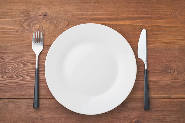 Clean empty white plate, fork and knife on brown wooden table, copy space, , top view.