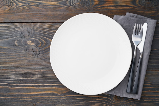 Clean empty white plate, fork and knife on dark rustic wooden table, copy space, , top view.