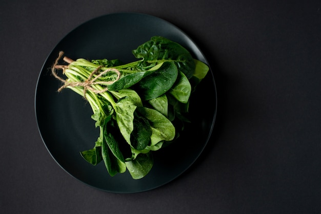 Clean food concept. Bunch of leaves of fresh organic spinach greens in a plate on a black background. Healthy detox spring-summer diet. Vegan Raw Food. Copy space.