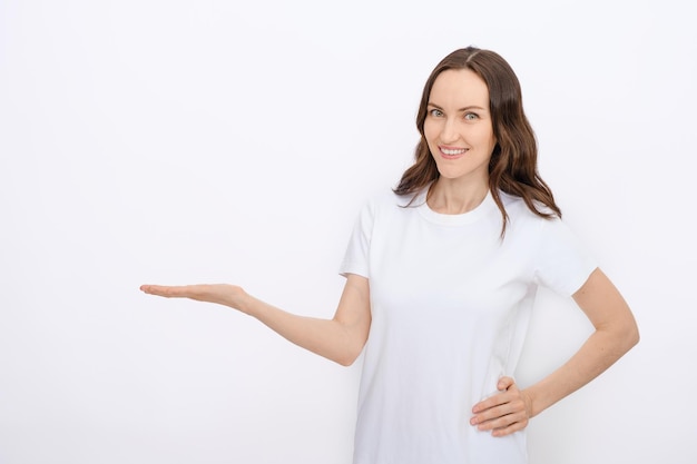 Clean minimal portrait of smiling brunette woman in white Tshirt on white background holds bent arm for advertising goods copy space template