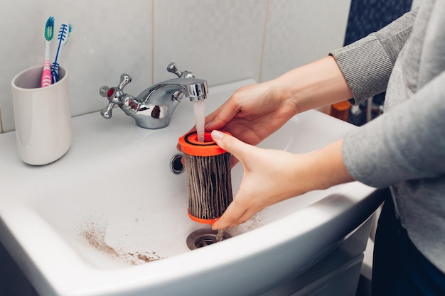 Cleaning vacuum cleaner filter woman washing dirty filter with water in bathroom sink at home